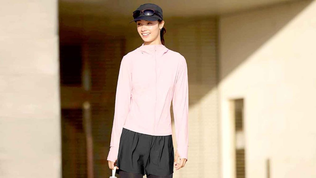 Model wanita bergaya kasual urban mengenakan jaket pelindung UV hitam dengan latar belakang arsitektur bangunan modern dan lapangan terbuka