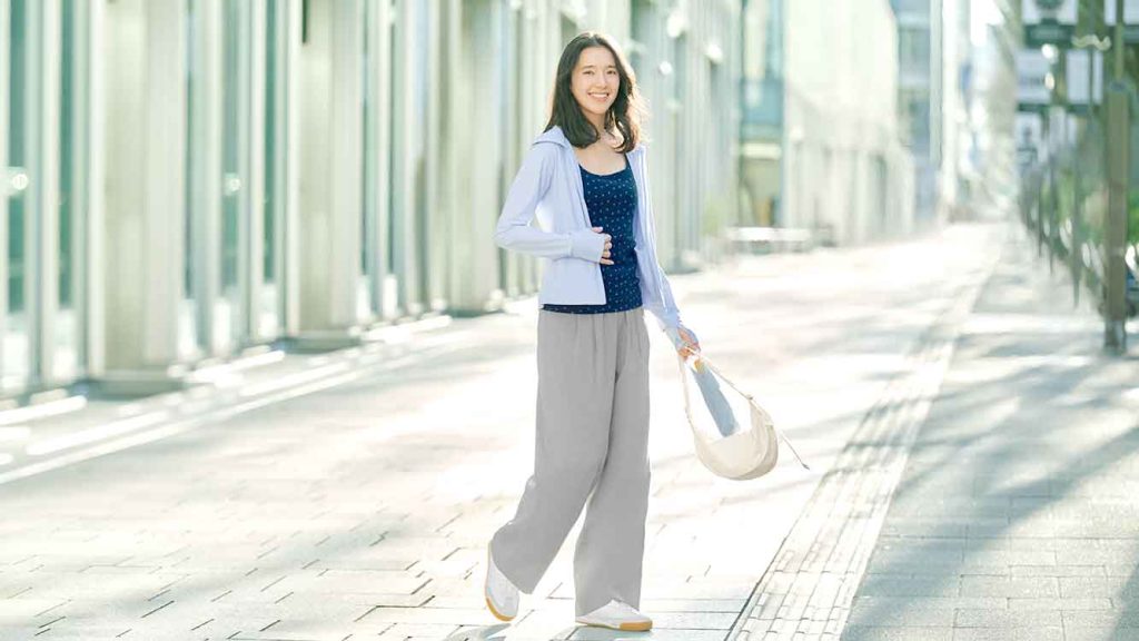Model wanita mengenakan jaket proteksi UV warna lilac dan celana bahan kerja di depan koridor gedung perkantoran modern